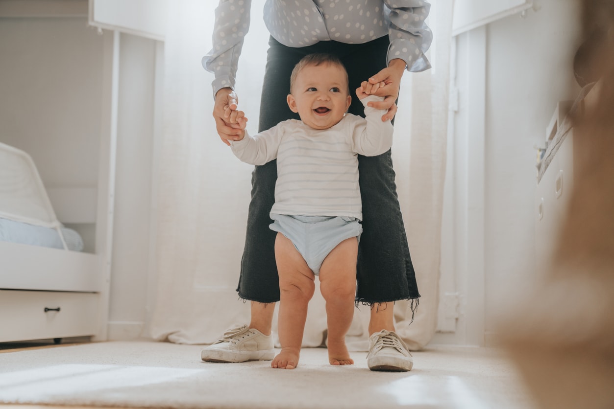 Woman stands behind the baby, holds his hands and helps him walk. The baby is smiling and happy.