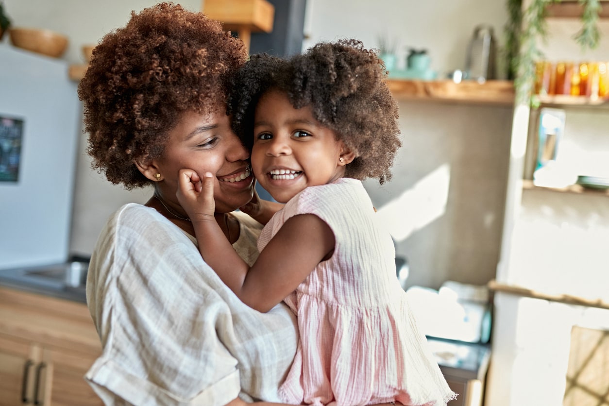 Partial side view of a 3 year old daughter smiling at the camera while her 26 year old mother holds her and expresses love and affection to her.