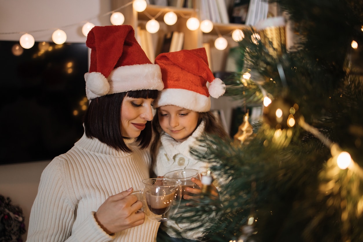 Close-up portrait of lovely family, beautiful young mother and her cute little daughter in warm knitted white sweaters drinking hot cocoa or chocolate from cups in front of decorated Christmas tree.