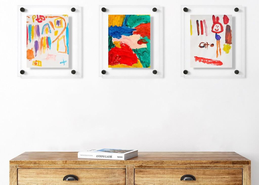Three colorful abstract paintings in white frames hang on a white wall above a wooden cabinet with two drawers. On the cabinet lies a book entitled ANDREAGIO. The paintings are characterized by bright colors and childlike drawings that are reminiscent of whimsical children's drawings.