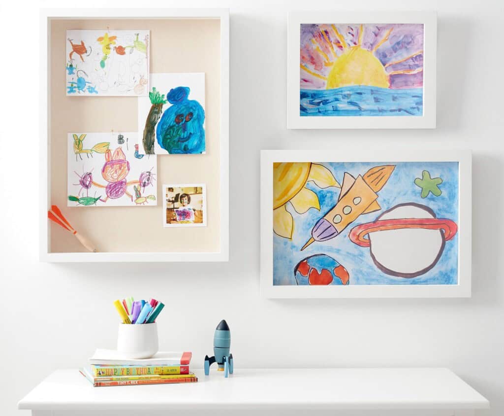 A white wall with framed children's pictures. One frame holds various small drawings pinned to a bulletin board. The other two frames show a colorful sunset and a space scene with a rocket. Below is a white desk with books, colored pencils in a cup, and a blue toy rocket.
