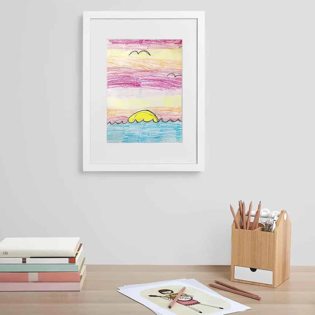 On a grey wall hangs a child's artwork, a coloured pencil drawing of a sunset over the sea in a white frame. Below it is a wooden desk with a stack of books, a drawing of a figure and a wooden holder with coloured pencils and scissors.