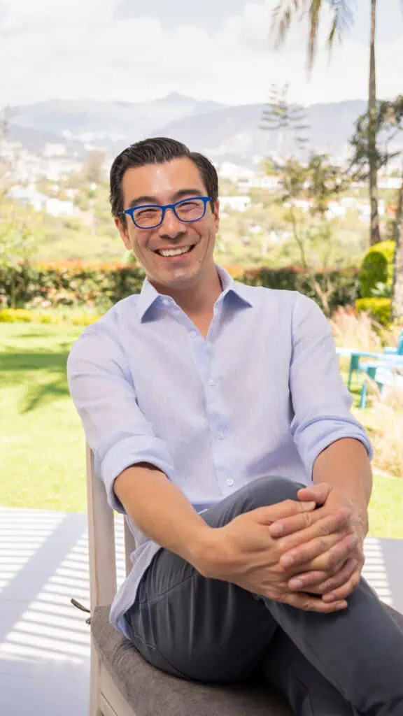 David Delisle sits on a chair outside in a light blue button-down shirt and dark pants. He is smiling, his legs are crossed and his hands are on his knees. He also wears blue glasses. The background shows a sunny landscape with lots of greenery and hills.