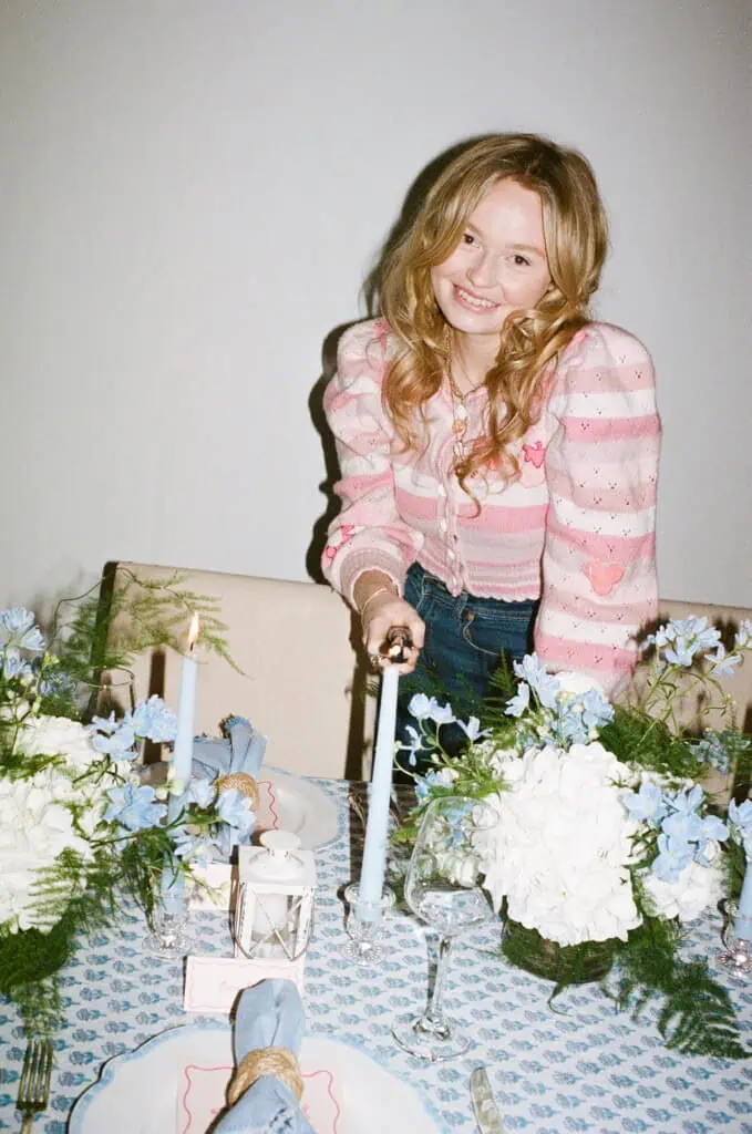 Cameron Forbes with long blonde hair, a pink striped sweater and jeans lights a blue candle on a dining table. The table is set with plates, glasses and floral arrangements with white and blue flowers. The ambience appears casual and decorative.