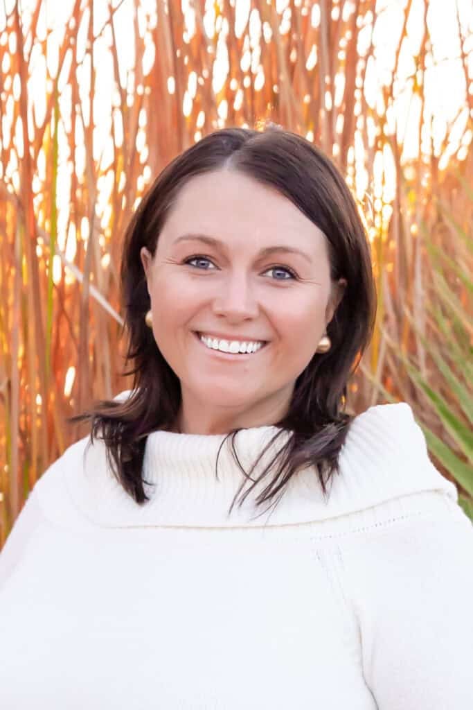 Jaryn Pederson smiles with dark hair while he stands in front of a high grass. They carry a white sweater and the sun shines brightly in the background.