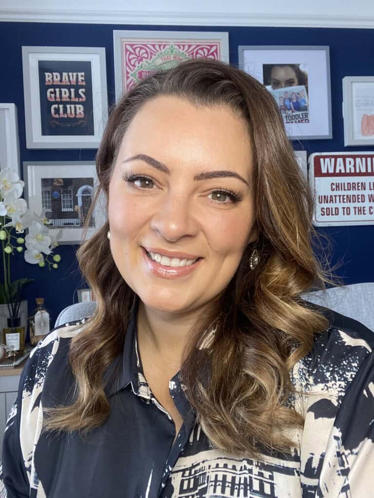 A woman with wavy brown hair smiles in a room with a dark blue wall with framed pictures and signs in the camera. She wears a patterned shirt and pearl earrings. White flowers are visible in the background.
