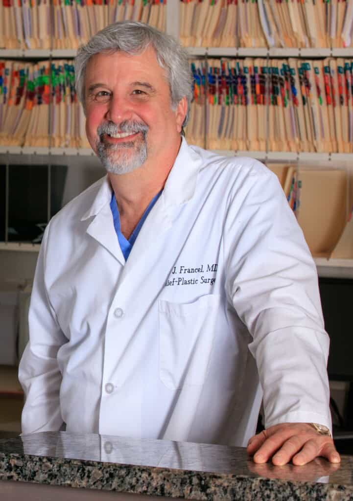 Dr. Thomas Francel with gray hair and a beard wears a white laboratory coat and stands on a granite counter with one hand. Behind him are shelves with organized folders.