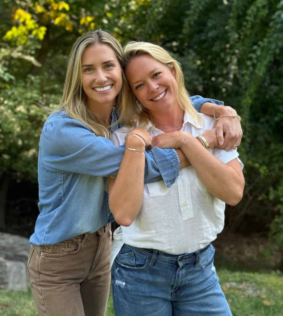 Dr. Jen and Bethany from the podcast podcast podcast in the imperial experience stand outdoors with green and smile in the camera. Dr. Jen hug Bethany from the side. Both carry long -sleeved shirts and jeans. The background includes trees and grass.