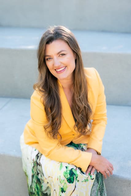 A woman with long brown hair sits on concrete steps wearing a yellow blazer and a patterned skirt. She is smiling and looking at the camera, her hands resting on her knees.